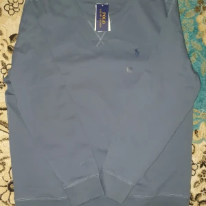 Blå långärmad tröja Polo Ralph Lauren oanvänd. - Snygg blå långärmad tröja från Polo Ralph Lauren med klassisk rund hals och diskret broderad logga på bröstet. Tröjan är tillverkad i mjuk bomull och har ribbade muddar vid ärmslut och nederkant. Perfekt för en chill och stilren look.