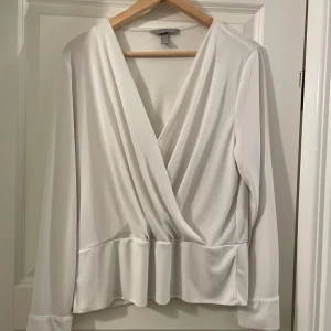 Vit omlottblus från H&M - Säljer en stilren vit omlottblus från H&M med lång ärm och djup v-ringning. Blusen har en snygg draperad effekt framtill och bredare fåll nedtill. 