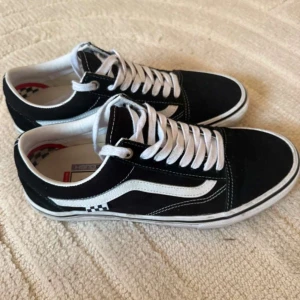 Svarta Vans Old Skool sneakers - Klassiska Vans Old Skool sneakers i svart med vit sidorand och vita snören. Skorna har checkerboard-detalj vid hälen, platt gummisula och ovandel i mocka och canvas. Perfekta för en streetstyle-look och passar till det mesta.