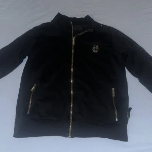 Svart Philipp Plein zip hoodie - Svart hoodie från Philipp Plein med hel dragkedja i guld, glittriga detaljer längs axlarna och stort logotryck på ryggen. Märkets emblem i metall framtill. Två fickor med dragkedja. Snygg och lyxig streetstil. Bra skick, 8/10!🔥