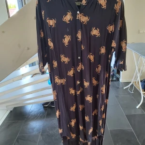 Mörk långklänning med krabbmönster - Säljer en cool mörk långklänning med unikt krabbmönster i beige. Klänningen har knappar framtill och trekvartsärm, vilket ger en avslappnad och trendig vibe. Perfekt för dig som gillar att sticka ut med roliga prints och bekväma plagg.