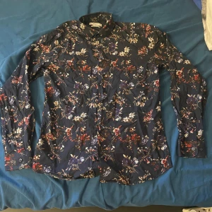 Blommig skjorta från Jack & Jones - Mörkblå skjorta från Jack & Jones med blommigt mönster i rött, vitt och lila. Slim fit-modell med långa ärmar och klassisk krage. Perfekt för dig som vill sticka ut med en snygg och färgstark look.