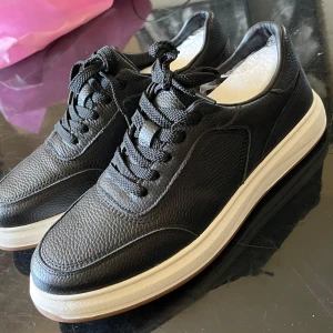 Svarta sneakers  - Stilrena svarta sneakers i grovpräglat skinn med klassisk snörning och vit platt sula. Skorna har rund tå och diskreta sömmar som ger en clean look. Perfekta för dig som gillar minimalistisk stil med en sportig touch.