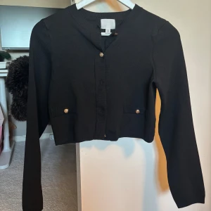 Svart kofta med guldknappar H&M - Stilren svart kofta från H&M med croppad passform och snygga guldknappar framtill samt på fickdetaljerna. Långärmad och ribbad vid kanterna, perfekt att slänga över en topp för en trendig look.