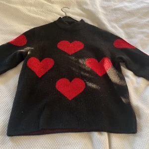 Svart stickad tröja med röda hjärtan - Cool svart stickad tröja med stora röda hjärtan över hela framsidan och ärmarna. Tröjan har en hög hals och långa ärmar. Supermysig och passar perfekt till jeans eller kjol.