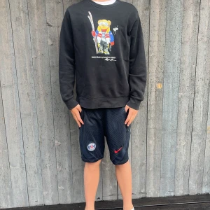 Svart Polo Bear tröja Ralph Lauren - Svart sweatshirt från Ralph Lauren med ikoniska Polo Bear-trycket framtill. Tröjan har rund hals, långa ärmar och en relaxed passform. Perfekt för dig som gillar streetstyle och vill sticka ut med en klassisk men lekfull look.