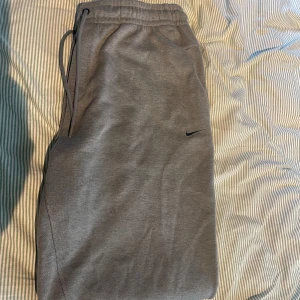 Grå mjukisbyxor från Nike Dri-Fit - Snygga grå mjukisbyxor från Nike med Dri-Fit-teknologi och standard fit. Byxorna har elastisk midja med snörning och diskret Nike-logga på benet. Perfekta för chill eller träning, tillverkade i mjukt och funktionellt material. Prislapp är kvar och har aldrig använts.