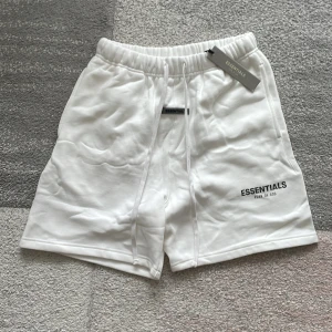 Vita Essentials shorts med snörning - Snygga vita shorts från Essentials Fear of God med elastisk midja och snörning framtill. Mjukt material, perfekt för chill dagar. Diskret logga på benet och bakficka för extra stilpoäng. Passar dig som gillar clean och enkel streetwear.