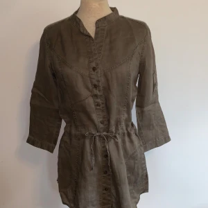 Militärgrön kortklänning/lång skjorta från MNG BASICS - Snygg militärgrön kortklänning alternativt lång skjorta från MNG BASICS med knappar framtill och knyt i midjan. Klänningen/skjortan har trekvartsärm som kan rullas upp och fästas, samt ståkrage och praktiska fickor. Lite militärtema. Lappen är tyvärr avklippt så vet inte vad det är för material.