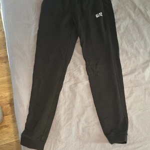 Svarta mjukisbyxor från EA7 Armani - Svarta mjukisbyxor från EA7 Emporio Armani med elastisk midja och smal passform. Byxorna har en diskret logga på ena benet och är tillverkade i mjukt bomullsmaterial. Perfekta för chill eller träning, med klassisk stil och bekväm känsla. Pris går att diskuteras.