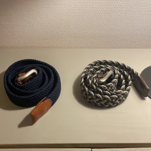 Två flätade bälten med läderdetaljer - Säljer två snygga flätade bälten med silverfärgat spänne och bruna läderdetaljer. Ett bälte är marinblått och det andra är randigt i beige, svart och grått. Perfekta accessoarer för att lyfta din outfit med en stilren touch.