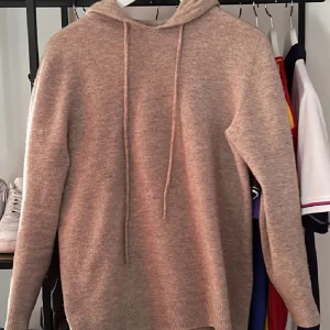 Beige stickad hoodie med huva - Mysig beige stickad hoodie med huva och snörning. Tröjan har lång ärm och ribbade muddar vid ärmslut och nederkant. Perfekt för lager-på-lager och chill dagar. Enkel och stilren design som passar till det mesta.