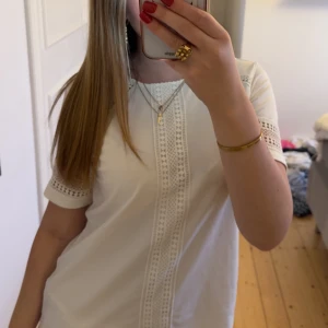 Vit t shirt med spetsdetaljer - Säljer en vit t shirt med snygga spetsdetaljer längs mitten och på ärmarna. T shirtenhar rund hals och korta ärmar med dekorativa mönster. Perfekt för dig som gillar en clean och fräsch look med lite extra detaljer.