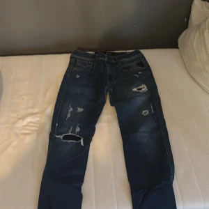Replay Anbass blå slitna jeans - Säljer ett par blå Replay Anbass jeans med snygga slitningar och hål på benen. Jeansen har klassisk femficksdesign, normal passform och är tillverkade i jeansmaterial. Perfekta för dig som gillar en lite ruffare streetstil. Kvitto kan visas vid intresse.