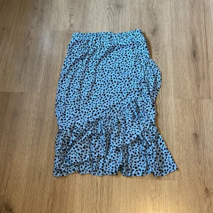 Blå kortkjol med volang och prickar - Supersöt blå kortkjol med svarta prickar och volangkant. Kjolen har omlottkänsla och är perfekt för dig som gillar lekfull stil. Lätt och luftig, passar perfekt till sneakers eller sandaler.