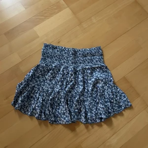 Blommig kortkjol från 157 - Supersöt blå kortkjol från 157 med vitt blommönster och volangkant. Kjolen har smockad midja som sitter skönt och ger en snygg passform. Perfekt för dig som gillar en feminin och chill stil.