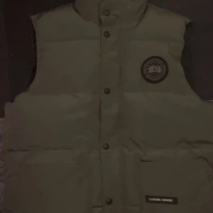 Grå väst från Canada Goose - Säljer en grå dunväst från Canada Goose med klassisk logga på bröstet. Västen har hög krage, tryckknappar framtill och två stora fickor. Perfekt för lager-på-lager och håller dig varm under kyliga dagar.