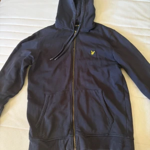 Marinblå hoodie från Lyle & Scott - Snygg marinblå hoodie från Lyle & Scott med dragkedja, huva och snörning. Klassisk gul logga på bröstet och två stora fickor framtill. Perfekt för chill dagar eller när du vill ha en enkel men stilren look.