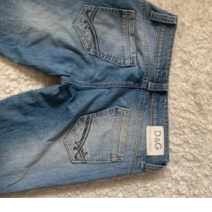 Blå jeans från D&G med broderade fickor - Snygga blå jeans från D&G med broderade detaljer på bakfickorna och klassisk femficksdesign. Jeansen har en rak passform och är tillverkade i mjukt denimtyg. Perfekta för dig som gillar en stilren men ändå unik look. 27:30