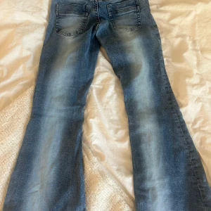 Blå bootcut jeans låg midja - Snygga blå jeans med bootcut och låg midja. Jeansen har klassisk femficksdesign och ljusa slitningar framtill och baktill för en cool, avslappnad look. Perfekta för dig som gillar retrovibbar och vill ha ett par jeans med lite extra attityd.