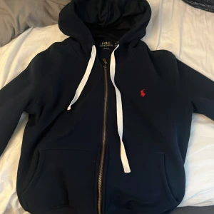 Mörkblå hoodie från Polo Ralph Lauren - Säljer en mörkblå hoodie från Polo Ralph Lauren med vit snörning och dragkedja. Klassisk röd broderad logga på bröstet och två fickor framtill. Priset kan diskuteras och mer bilder kan läggas till vid efterfrågan