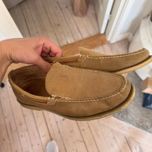 Beige mockaloafers från Polo Ralph Lauren - Snygga loafers i beige mocka från Polo Ralph Lauren med broderad logga på ovandelen. Skorna har klassisk seglarskodesign med ljus sula och kontrastsömmar. Perfekta för en avslappnad men stilren look.