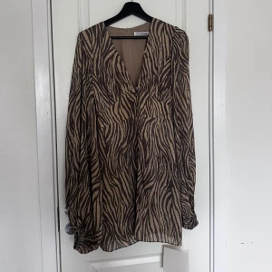 Zebramönstrad miniklänning från Adoore - Säljer en beige och brun zebramönstrad miniklänning från Adoore med v-ringning och knappar framtill. Klänningen har långa ärmar med lätt puff och är tillverkad i ett tunt, flowy material som ger en snygg siluett. 