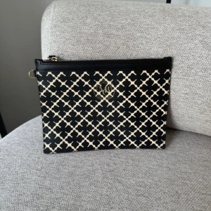 Svart och beige kuvertväska från By Malene Birger - Stilren kuvertväska från By Malene Birger i svart och beige med ikoniskt grafiskt mönster och guldfärgad logga framtill. Väskan har dragkedja upptill och är tillverkad i skinnliknande material. Perfekt för att bära det viktigaste med stil.