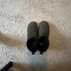 Svarta UGG boots i mocka - Säljer ett par klassiska svarta UGG boots i mocka med mjukt foder och platt sula. Skorna har rund tå och dragflik baktill för enkel påtagning. Perfekta för kyliga dagar och riktigt sköna att ha på sig.