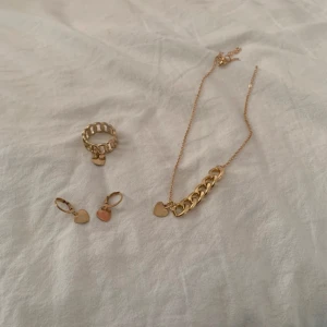 Guldigt smyckeset med hjärtan - Snyggt smyckeset i guld med chunky ring, halsband och örhängen, alla med små hjärtanhängen. Perfekt för dig som gillar detaljer och vill ha ett matchande set. Kedjan på halsbandet är delvis grov och örhängena är små hoops med hjärtberlocker.
