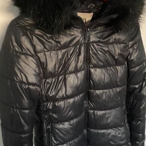 Svart pufferjacka med fur - Snygg svart pufferjacka med glansig finish och huva med fuskpäls. Jackan har dragkedja framtill och två fickor med dragkedja. Perfekt för kalla dagar när du vill hålla stilen och värmen.