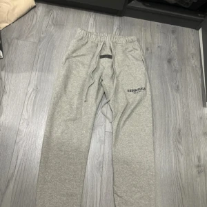 Grå Essentials Fear of God mjukisbyxor - Snygga grå mjukisbyxor från Essentials Fear of God med logga tryckt på benet och en svart patch framtill. Byxorna har elastisk midja med snörning och är gjorda i mjukt bomullsmaterial. Perfekta för chill eller streetwear-stil.