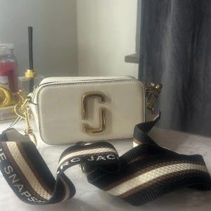 Vit axelväska från Marc Jacobs - Snygg vit axelväska från Marc Jacobs med guldiga detaljer och bred, randig axelrem i svart, brunt och beige. Väskan har två dragkedjor upptill, ett stort guldigt logomärke framtill och en praktisk ficka på baksidan. Perfekt för att lyfta vilken outfit som helst.