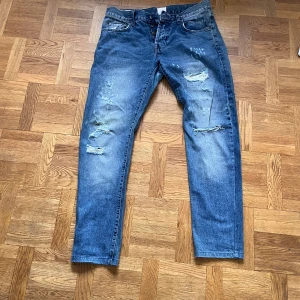 Blå slitna jeansbyxor Ruffle & bow 32/34 - Säljer ett par blå jeansbyxor från Ruffle & bow i storlek 32/34. Jeansen har slitna detaljer och hål på både fram- och baksida, klassisk femficksmodell och knappgylf. Materialet är jeans och passformen är rak med normal midja.