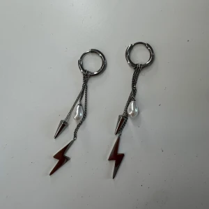 Örhängen med blixt, pärla och spike - Säljer ett par coola örhängen i silverfärg med ring, hängande blixt, konformad spike och en vit pärldroppe. Kedjorna ger en edgy vibe och kombon av metall och pärla gör dem unika. Perfekta för dig som vill sticka ut med din stil.