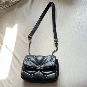 Svart axelväska med kedja - Snygg svart axelväska i skinnimitation med coola guldfärgade nitar. Väskan har en bred axelrem i kombination med en kedjedetalj i metall. Perfekt storlek för det viktigaste och med ett fack på insidan för extra organisering 🖤