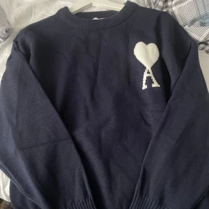 Mörkblå stickad tröja från Ami - Säljer en mörkblå stickad tröja från Ami Alexandre Mattiussi med vit hjärtlogga och A på bröstet. Tröjan har rund halsringning, långa ärmar och ribbade muddar. Perfekt för dig som gillar stilrena och trendiga plagg.