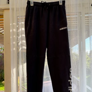 Svarta mjukisbyxor från Adidas - Svarta mjukisbyxor från Adidas med stor logga längs ena benet i grått och vitt mönster. Byxorna har resår och snörning i midjan samt sidofickor. Perfekta för chill eller träning, riktigt sköna och stilrena.