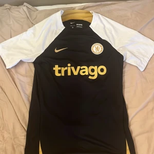 Chelsea svart och vit fotbollströja & shorts Nike. Storlek S - Chelsea FC fotbollströja från Nike i slim fit Dri-FIT material. Svart kropp med vita korta ärmar och guldiga detaljer på krage, logga och tryck. Tröjan har Chelsea-emblem på bröstet och trivago-tryck framtill. Perfekt för träning eller match.