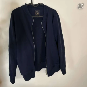 Marinblå ziptröja från Marine Sailing Club - Snygg marinblå ziptröja från Marine Sailing Club med broderad logga i nacken. Tröjan har långa ärmar, ribbade muddar och dragkedja framtill. Perfekt för dig som gillar en klassisk och stilren look. Passar till både jeans och chinos.