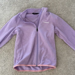 Ljuslila fleecejacka från Everest - Supermjuk ljuslila fleecejacka från Everest med hel dragkedja och diskret logga på bröstet. Jackan har långa ärmar och rosa kantband nedtill för en snygg kontrast. Perfekt för kyliga dagar eller som lager under jackan.