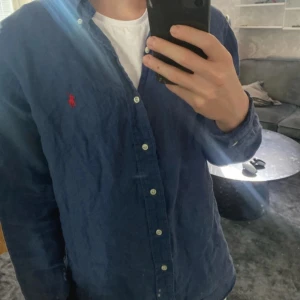 Blå skjorta från Ralph Lauren - Säljer en klassisk blå skjorta från Ralph Lauren med röd broderad logga på bröstet. Skjortan har knappar hela vägen och lång ärm, perfekt för en stilren look. Tillverkad i mjuk bomull som känns skön mot huden.