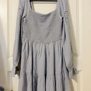 Ljusgrå broderad klänning  - Supersöt ljusgrå klänning  med smockad överdel och volangkjol. Långa ärmar med broderade detaljer och hålmönster ger en unik vibe. Perfekt fyrkantig ringning och skön passform. Materialet känns mjukt och luftigt, idealiskt för vår och sommar.