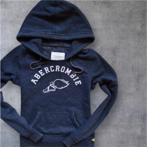 Mörkblå hoodie från Abercrombie - Mörkblå hoodie från Abercrombie & Fitch med vit logga och snörning i huvan. Tröjan har en klassisk känguruficka och långa ärmar. Perfekt för en avslappnad och cool stil.