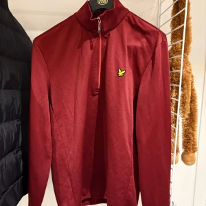 Vinröd half zip tröja från Lyle & Scott - Vinröd långärmad tröja från Lyle & Scott med half zip och ståkrage. Tröjan har det klassiska gula fågel-loggan på bröstet och är gjord i mjuk silikon . Perfekt för dig som gillar stilren och sportig look.