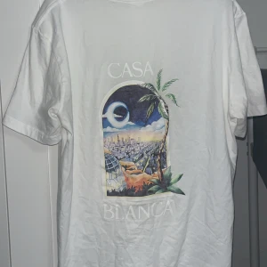 Vit Casa Blanca t-shirt med tryck - Vit t-shirt från Casa Blanca med färgstarkt grafiskt tryck på ryggen och mindre tryck på bröstet. Motivet visar en stad, palmer och måne i en cool illustration. T-shirten har rund hals och korta ärmar, perfekt för dig som gillar streetwear med unik design. Storlek L men passar M också. Pris kan diskuteras! 