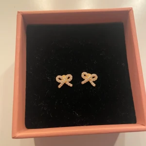 Guldiga rosettörhängen med pärlor - Supersöta örhängen i form av små rosetter, gjorda i guldfärgad metall och dekorerade med glittrande pärlor. Perfekta för dig som gillar detaljer och vill ha något extra till din look. Säljs i originalask.