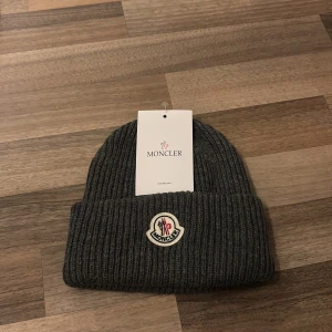 Mörkgrå ribbad Moncler mössa - Snygg mörkgrå mössa från Moncler med ribbad stickning och klassisk Moncler-logga framtill. Mössan har uppvikt kant och är tillverkad i mjuk ull som håller dig varm. Perfekt för dig som vill ha en clean och exklusiv look under kyliga dagar. Aldrig använt. Skannar inte. Pris sitter inte fast i sten. Inget kvitto