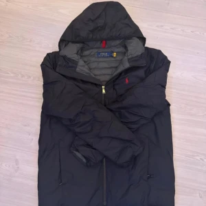 Ralph lauren jacka - Svart dunjacka från Polo Ralph Lauren med huva och dragkedja. Jackan har den klassiska röda Polo-loggan broderad på bröstet och är lätt att packa ner i medföljande påse. Perfekt för kalla dagar och enkel att ta med sig. Snygg och stilren design.