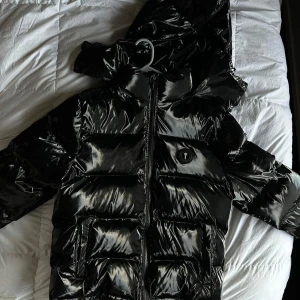 Svart pufferjacka från Trapstar - Säljer en svart, glansig pufferjacka från Trapstar med stor huva och fet logga på ryggen. Jackan har dragkedja framtill, sidofickor och en rund Trapstar-patch på bröstet. Perfekt för dig som vill sticka ut med streetstyle-vibe under kalla dagar.